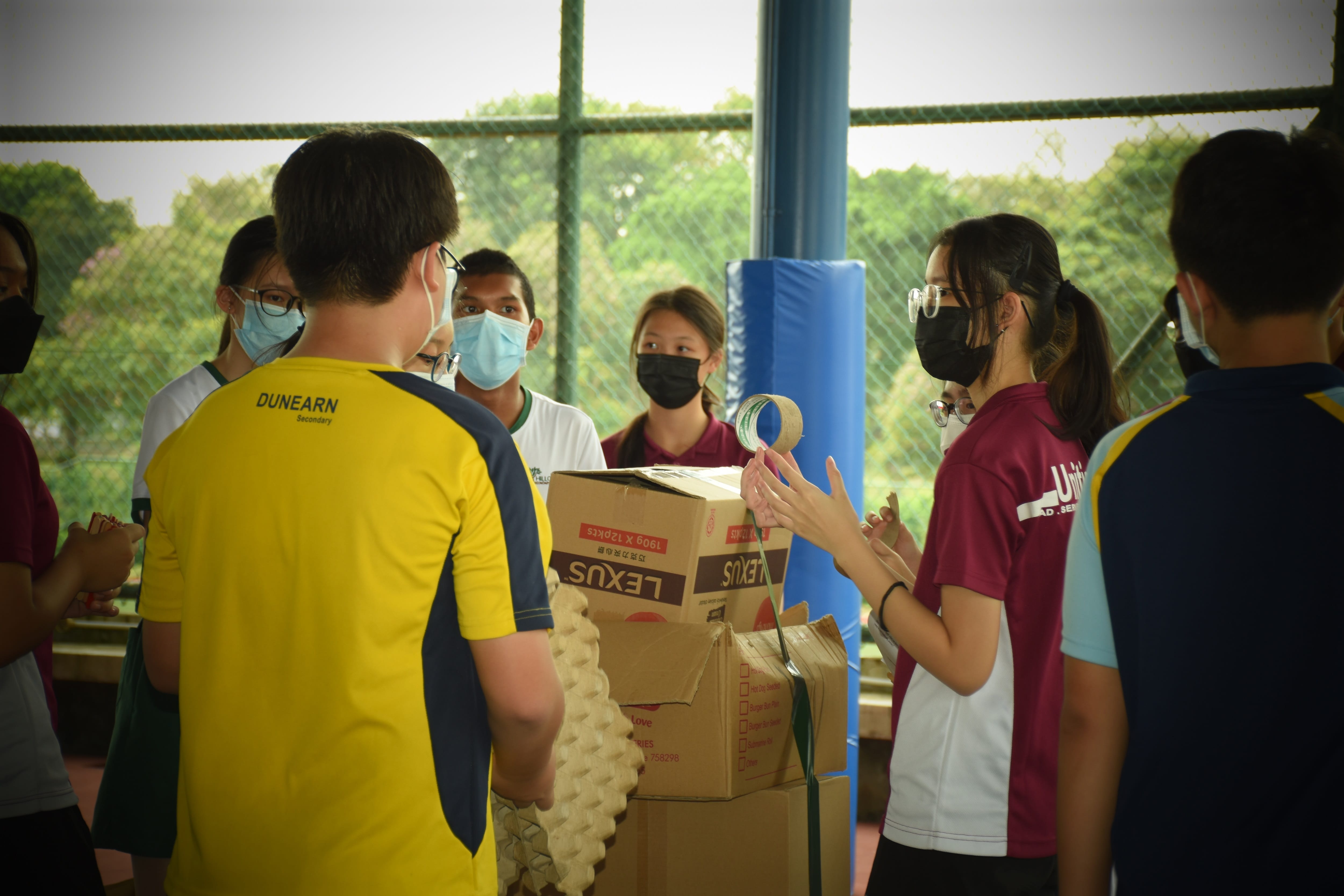 Students in school uniforms and face masks gather around cardboard boxes and a roll of tape outdoors.
